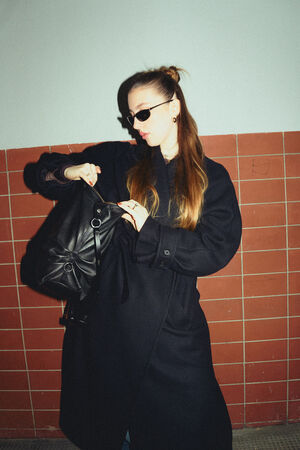 A female-presenting person is wearing a long black coat, sunglasses and holding the “DELA EDEN ed.1” bag in pure black. She is reaching into the bag and has her hair tied back.
