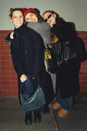 Three female-presenting people stand together smiling in front of a wall with red tiles. The person on the left is holding a blue, shiny bag named "DELA YIN ed.1".