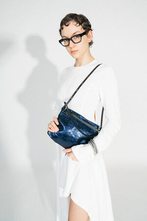 A MOONLIT ed.1 bag in the color Crackled Deep Navy, worn diagonally over the shoulder. The metallic, deep blue color of the bag stands out against the wearer's simple white dress. The bag has a smooth, slightly structured surface with a black shoulder strap.