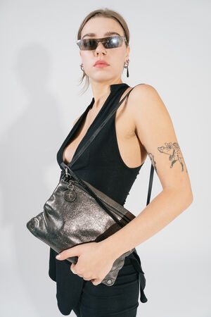 A person with a modern look carries a metallic dark anthracite-colored leather bag across the body. The bag has a shimmering effect, a visible zipper and a round leather tag. The person is wearing a black sleeveless top, sunglasses and has an eye-catching tattoo on their arm.