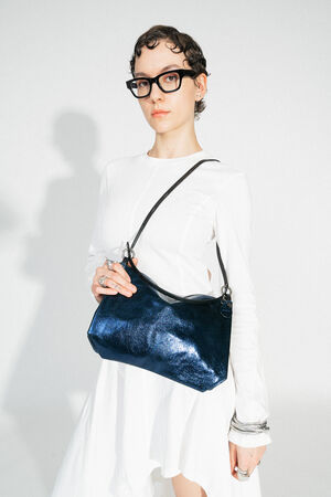 INA KENT ORNAMENT ed.2 crackled deep navy (sideways, worn over the shoulder): A dark blue bag with a shiny, textured surface is worn over the shoulder by a person in a white dress. The elegant and modern style of the bag complements the outfit.