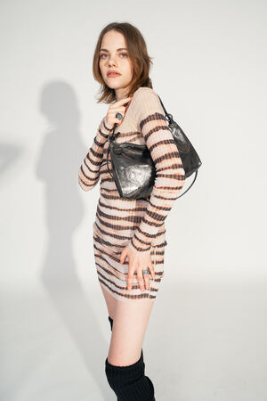 A female model carries the INA KENT ORNAMENT ed.2 bag in crackled graphite leather on her shoulder. She wears a striped, figure-hugging dress and looks directly into the camera. The bag with its shimmering metallic finish and sleek design is the focus of the picture.