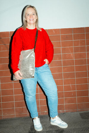 A female-presenting person is wearing a red sweater, blue jeans and white sneakers. She is carrying the “SHER ed.1” bag in crackled anthra over her shoulder.