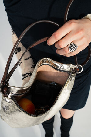 Die metallic-silberne MOONLIT ed.1 Tasche von INA KENT wird crossbody getragen und offenbart ihren Inhalt – eine Geldbörse, Schlüssel, ein Handy und ein Apfel. Das weiche Leder schimmert, ergänzt durch braune Lederriemen und einen charakteristischen Metallanhänger.