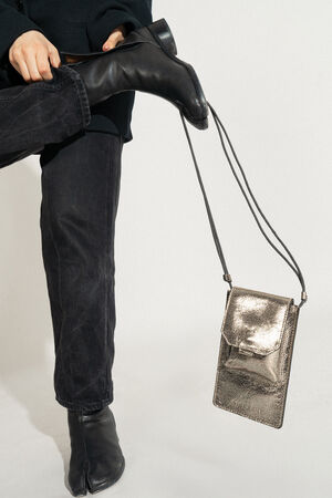 Die Person trägt ein elegantes schwarzes Outfit und Stiefel und rückt sorgfältig einen Schuh zurecht, während an ihrem Fuß eine elegante metallisch-silberne Tasche von INA KENT baumelt.