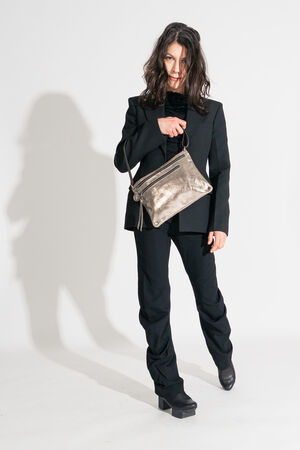 Person in a sleek black suit and platform shoes strikes a pose, showcasing an INA KENT metallic purse against a plain white background.