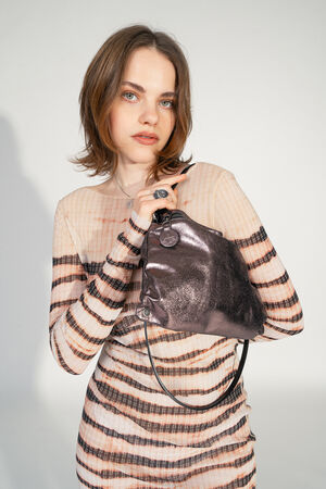 Woman in a chic striped dress holds a glossy metallic bag from INA KENT, standing elegantly against a neutral background.