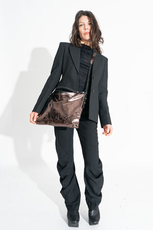 Person in a black suit and platform shoes showcases an exquisite INA KENT handbag, its shiny brown finish striking against the white background.