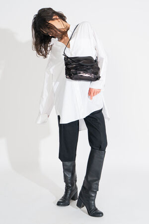 Person wearing an oversized white shirt, black pants, and knee-high boots, with a stylish black INA KENT shoulder bag, poses against a plain background.