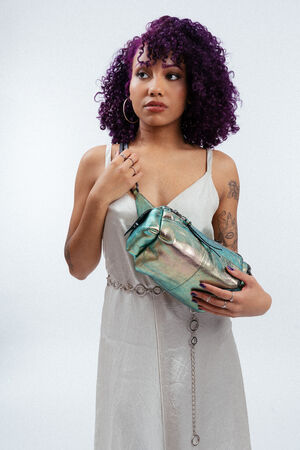 Frau mit lockigem lila Haar in einem weißen Kleid hält eine glänzend grüne INA KENT-Handtasche und schaut zur Seite; auf ihren Armen sind Tattoos sichtbar.