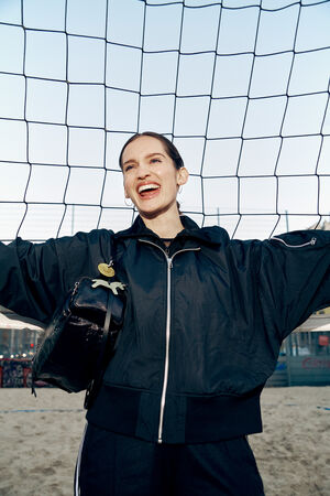 A person wearing a black jacket and an INA KENT backpack stands in front of a volleyball net on a sandy court, smiling with arms outstretched.