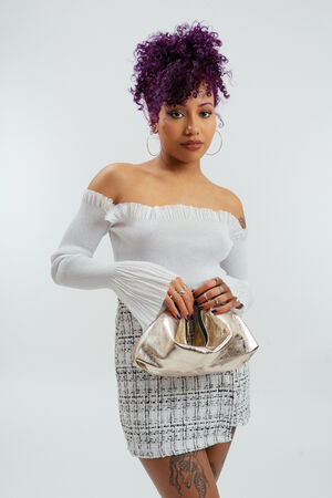 Frau mit lila Locken, schulterfreiem weißen Top und kariertem Rock, hält eine metallische Clutch von INA KENT und steht vor einem schlichten hellen Hintergrund.