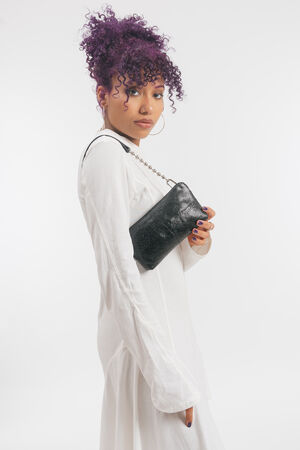 Model mit lila Haaren in weißem Kleid mit schwarz glänzenden Schultertasche 