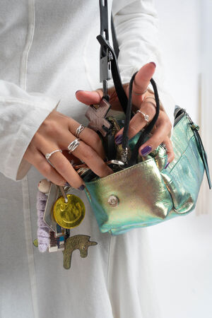 Eine Person in einem weißen Outfit hält eine schillernde INA KENT-Handtasche, die mit mehreren Schlüsseln und einem Einhorn-Schlüsselanhänger verziert ist, und greift mit perfekt manikürten Händen hinein.