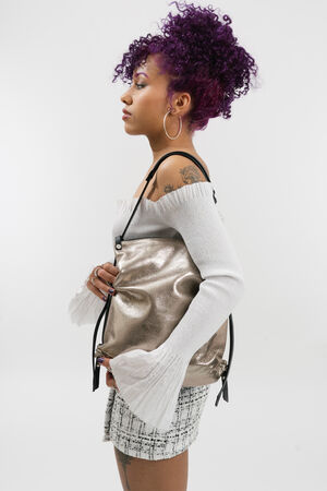 A woman with curly purple hair, wearing a white off-shoulder top and plaid skirt, poses against a white background with an INA KENT metallic gold bag slung stylishly over her shoulder.