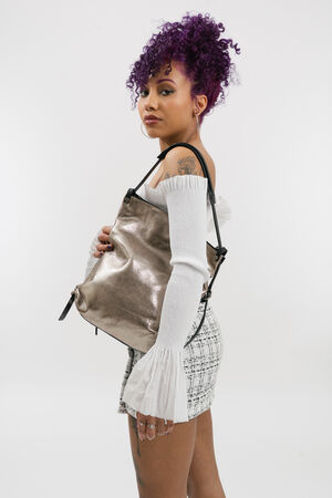 A woman with curly purple hair poses sideways, wearing a white off-shoulder top, patterned skirt, and holding a metallic INA KENT shoulder bag.
