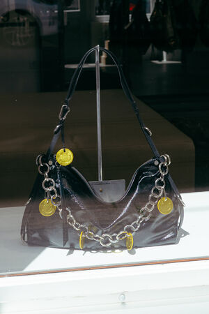 An INA KENT black patent leather handbag with a thick silver chain and large gold coin charms is displayed in a shop window.