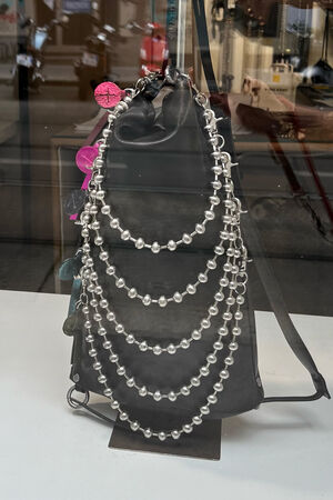 An INA KENT black leather backpack is displayed in a store window, decorated with five strands of silver beads and several colorful keychains on one side.