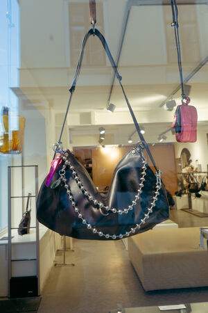 An INA KENT black leather handbag with a chain detail and a pink keychain is displayed in a store window with other bags in the background.