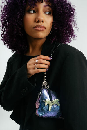 Eine Frau mit lockigem lila Haar und einem schwarzen Blazer hält eine kleine metallisch-lila Handtasche von INA KENT mit Einhorn-Design und einem Schlüsselanhänger.