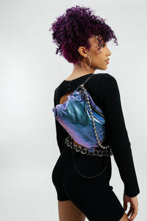 Person mit lockigem lila Haar, schwarzem Outfit und großen Creolen, die eine metallisch lila Tasche von INA KENT mit Kettenriemen über der Schulter trägt.