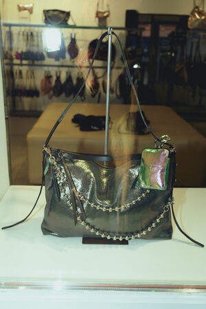 A shiny black INA KENT handbag with silver chain detailing and a small attached pouch is displayed in a glass case, with other handbags visible on shelves in the background.