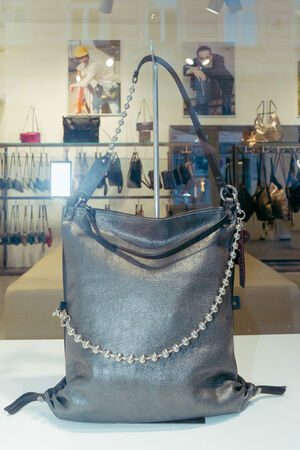 An INA KENT metallic shoulder bag with a beaded chain detail is displayed on a stand in a store, with other bags and wall art visible in the background.