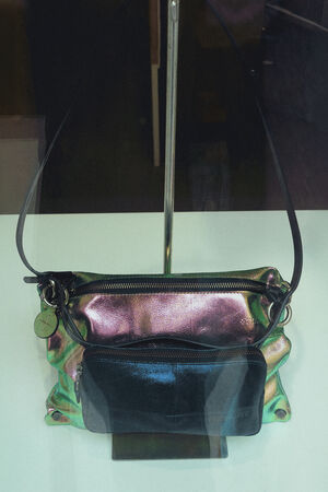 A shiny, iridescent INA KENT handbag with a black front pocket and a shoulder strap is displayed on a white surface.