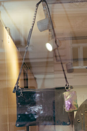 A black leather shoulder bag by INA KENT with a silver strap and a small iridescent pouch is displayed in a store window.