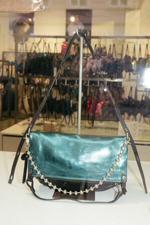A metallic teal INA KENT handbag with a decorative chain strap is displayed on a white surface, with shelves of other handbags visible in the background.