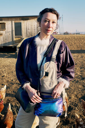 A woman wearing a purple jacket with fur trim and overalls stands outside on a farm near chickens, holding an INA KENT bag, with a wooden coop and clear sky in the background.