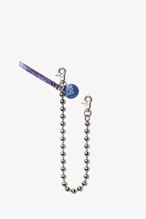 Eine Metallkugelkette mit zwei Karabinerverschlüssen, einem blauen runden Anhänger und einem glitzernden lila INA KENT-Etikett auf schlichtem weißem Hintergrund.