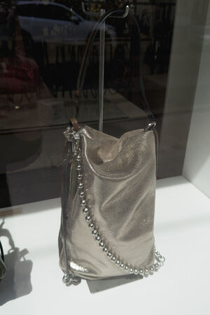 A metallic silver INA KENT handbag with a pearl-accented strap is displayed on a white surface in a store window.