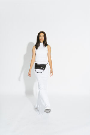 Eine Frau in einem ärmellosen weißen Kleid und einer schwarzen INA KENT-Gürteltasche steht vor einem schlichten weißen Hintergrund.