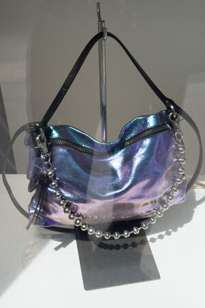 A metallic iridescent INA KENT handbag with black straps and a silver beaded handle is displayed on a stand behind glass.