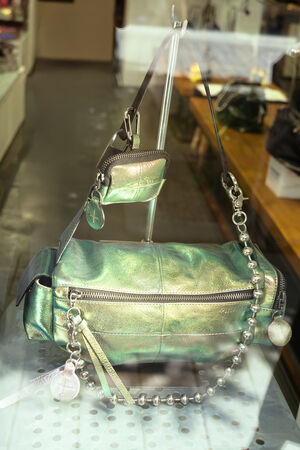 A metallic green INA KENT handbag with a front zipper, attached coin pouch, decorative beads, and keychain is on display behind a glass window.