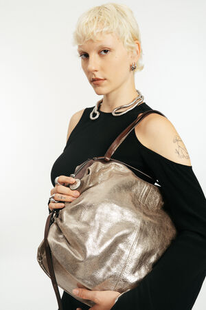 Person mit kurzen blonden Haaren, die ein schwarzes Oberteil trägt, hält eine große metallische INA KENT-Tasche mit braunen Trägern vor einem schlichten weißen Hintergrund.