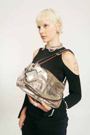 Person mit kurzen blonden Haaren, die ein schwarzes ausgeschnittenes Oberteil trägt, hält eine INA KENT Metallic-Silber-Handtasche über ihrer Schulter vor einem unifarbenen Hintergrund.