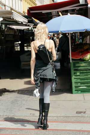 A person with blonde hair, wearing a black dress, gray tights, black boots, and carrying an INA KENT bag, walks through an outdoor market area with stalls and umbrellas.