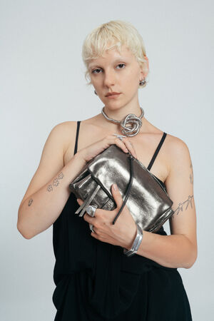 Person mit kurzen blonden Haaren und Tattoos hält eine INA KENT Metallic-Clutch-Tasche, trägt ein schwarzes ärmelloses Outfit und Silberschmuck, steht vor einem schlichten hellen Hintergrund.