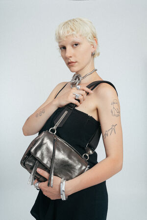 Person mit kurzen blonden Haaren in einem schwarzen Kleid hält eine INA KENT Metallic-Handtasche, zeigt sichtbare Tattoos und Silberschmuck, steht vor einem schlichten Hintergrund.