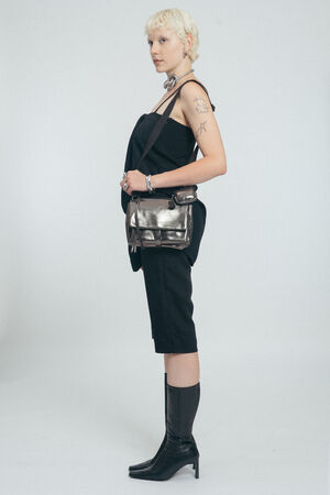 Eine Person mit kurzen blonden Haaren steht im Profil und trägt ein schwarzes Outfit, kniehohe schwarze Stiefel, silberne Accessoires und eine metallene INA KENT-Umhängetasche vor einem schlichten Hintergrund.