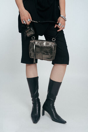 Person in knielangen schwarzen Shorts, schwarzen kniehohen Stiefeln und mit einer INA KENT Metallhandtasche und Schlüsseln in der Hand, wobei silberne Ringe und Armbänder zu sehen sind. Der Oberkörper ist größtenteils außerhalb des Bildausschnitts.
