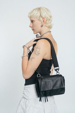 Eine Person mit kurzen blonden Haaren, bekleidet mit einem schwarzen Tanktop und einem weißen Rock, trägt eine schwarze INA KENT-Umhängetasche und hat sichtbare Tätowierungen auf dem Arm.