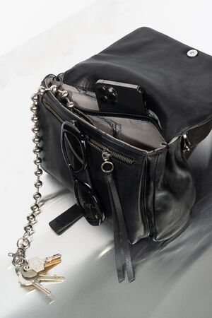Eine schwarze Ledertasche von INA KENT liegt offen auf einer hellen Unterlage und enthält ein Telefon, eine Sonnenbrille und eine Kette mit Schlüsseln daran.