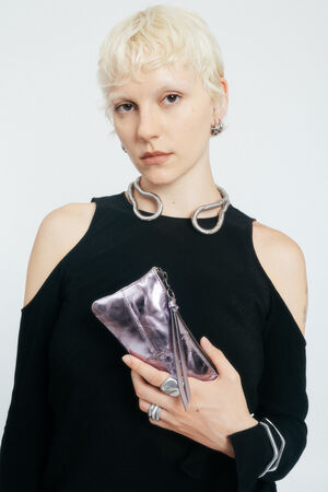 Person with short platinum blonde hair wears a black cold-shoulder top, silver jewelry, and holds an INA KENT shiny metallic purple clutch against a plain white background.