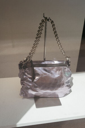 Eine silbermetallicfarbene INA KENT-Handtasche mit einem klobigen Kettenriemen und einem Reißverschluss an der Vorderseite wird auf einem Metallständer vor einem schlichten Hintergrund präsentiert.