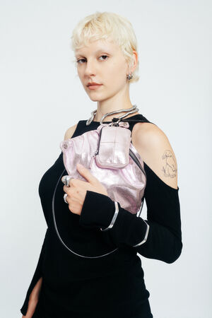 A person with short blond hair and a tattoo on their upper arm holds a shiny pink INA KENT bag while wearing a black cut-out dress.