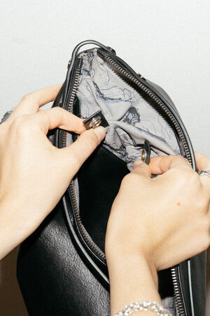 Die Hände einer Person reparieren das Innenfutter einer schwarzen INA KENT-Ledertasche und halten Stoff und einen Metallring in der Nähe eines offenen Reißverschlusses.