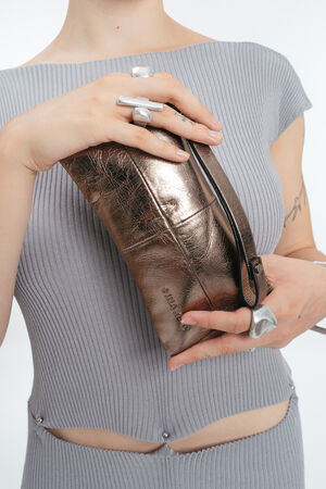 Eine Person in einem gerippten, hellgrauen Outfit hält eine INA KENT Metallic-Bronze-Armbandtasche; sie trägt klobige Silberringe an ihren Fingern.